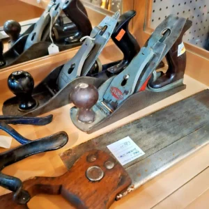 Antique woodworking hand planes and a Disston Steel handsaw with wooden handles arranged for sale on a shelf.