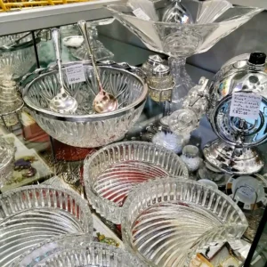 Display case featuring elegant crystal serving bowls, silver-plated salad servers, and a chrome "Rum Barrel" dispenser.