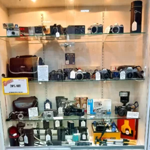 Glass display cabinet featuring a collection of vintage SLR, film, and digital cameras from Nikon, Pentax, and Canon.