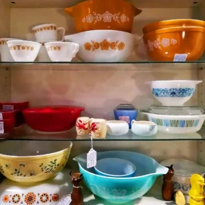Collection of vintage Pyrex and Corningware mixing bowls in various floral patterns and colors on glass shelves.