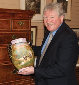 Henry Jones showing off some of Roadshow Antiques finest ceramics in a suit and tie.