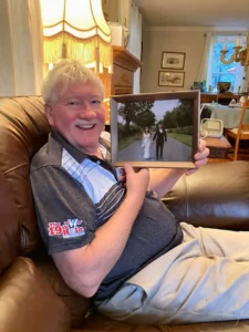 Henry Jones smiling while holding a framed wedding photograph in his living room.
