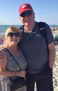 Henry Jones and Jackie posing together on a sandy beach during a bright, sunny day.