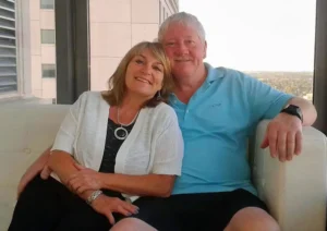 Henry Jones and Jackie relaxing together on a white sofa with a high-rise city view in the background.