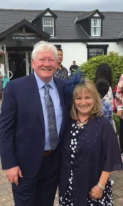 Henry Jones in a suit and Jackie in a navy dress and fascinator, posing outside the historic Gretna Green building.