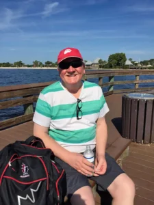 Henry Jones seated on a wooden pier by the water wearing a green striped shirt and red cap.