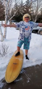 A humorous photo of Henry Jones in a Hawaiian shirt and blonde wig, posing with a surfboard in a snowy driveway.