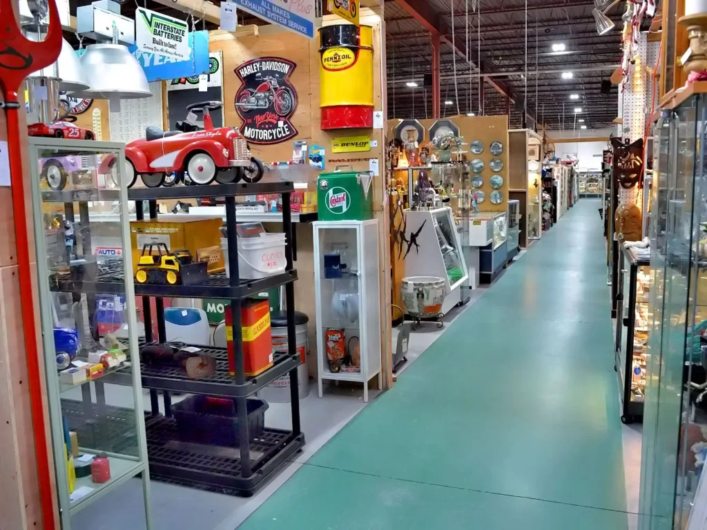 Roadshow Antiques Mall aisle lined with vendor booths featuring a vintage red pedal car, antique advertising signs including Harley-Davidson and Pennzoil, retro oil cans, vintage toys, and glass display cases stretching into the distance