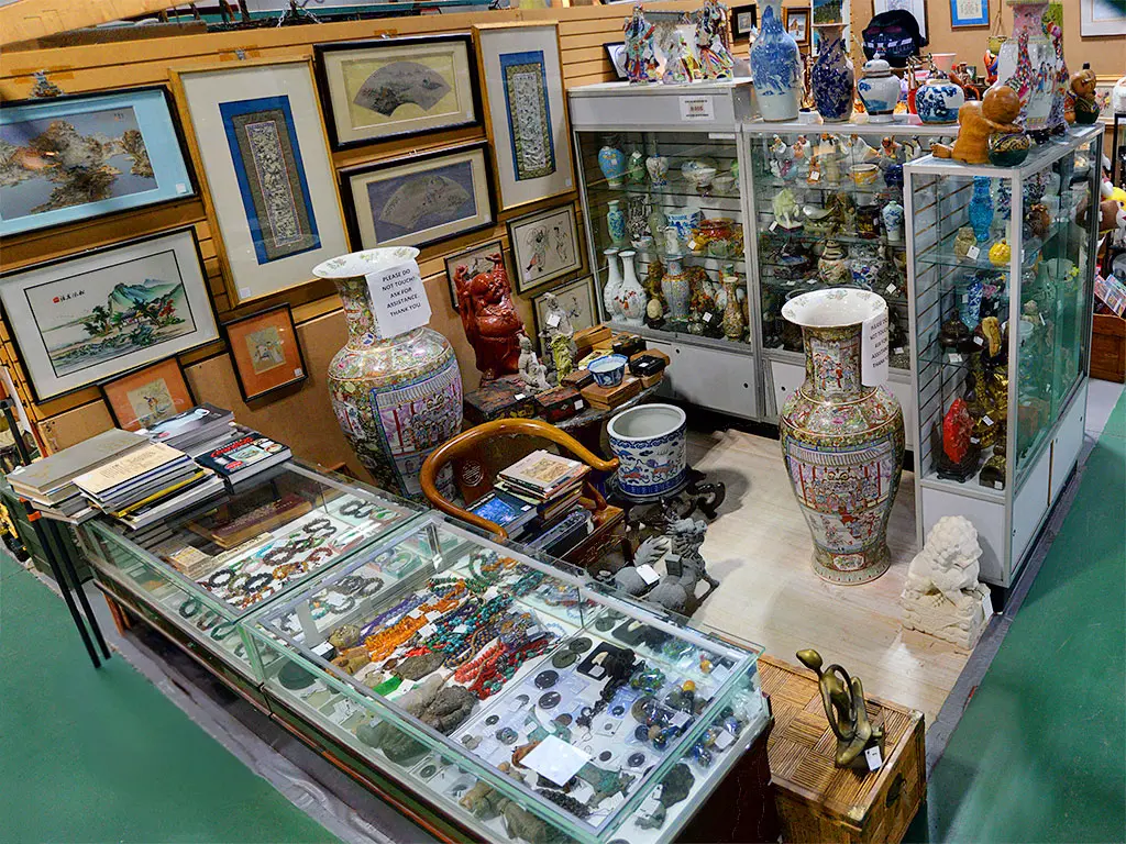 Asian antiques booth at Roadshow Antiques Mall featuring large Chinese porcelain vases, framed silk paintings, jade and gemstone jewelry in glass display cases, foo dog sculptures, and an extensive collection of decorative figurines