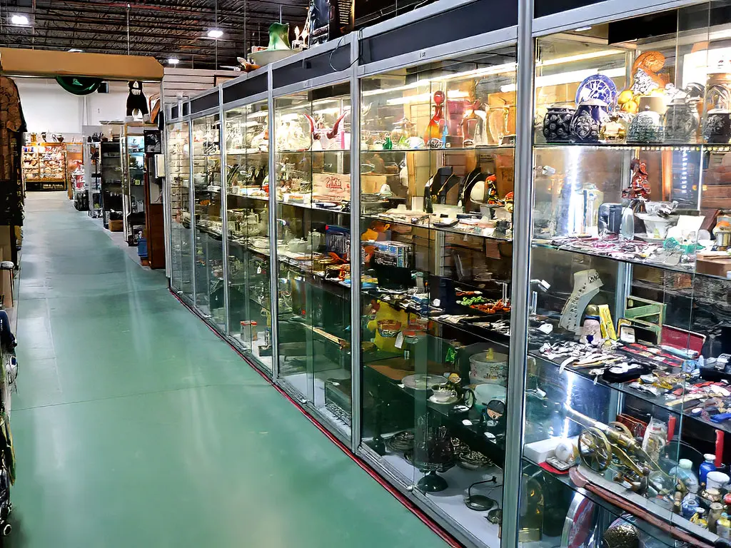 Long row of illuminated glass display cases at Roadshow Antiques Mall packed with small collectibles, jewelry, art glass, silverware, and vintage curiosities stretching along the aisle