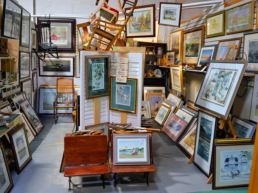 Densely packed art booth at Roadshow Antiques Mall featuring hundreds of framed prints, original paintings, and limited edition artwork covering the walls and floor, with antique rocking chairs and a vintage school bench