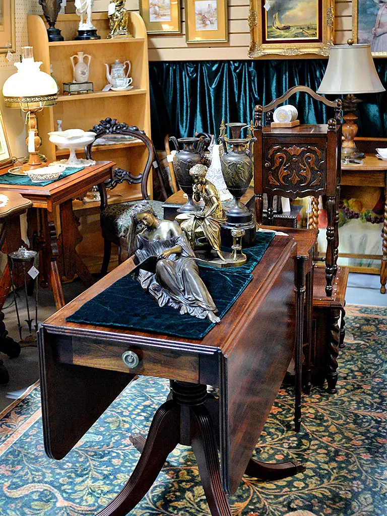 Antique mahogany drop-leaf table displayed with bronze figurines and classical urns at Roadshow Antiques Mall, surrounded by ornate carved furniture, framed artwork, and vintage lighting