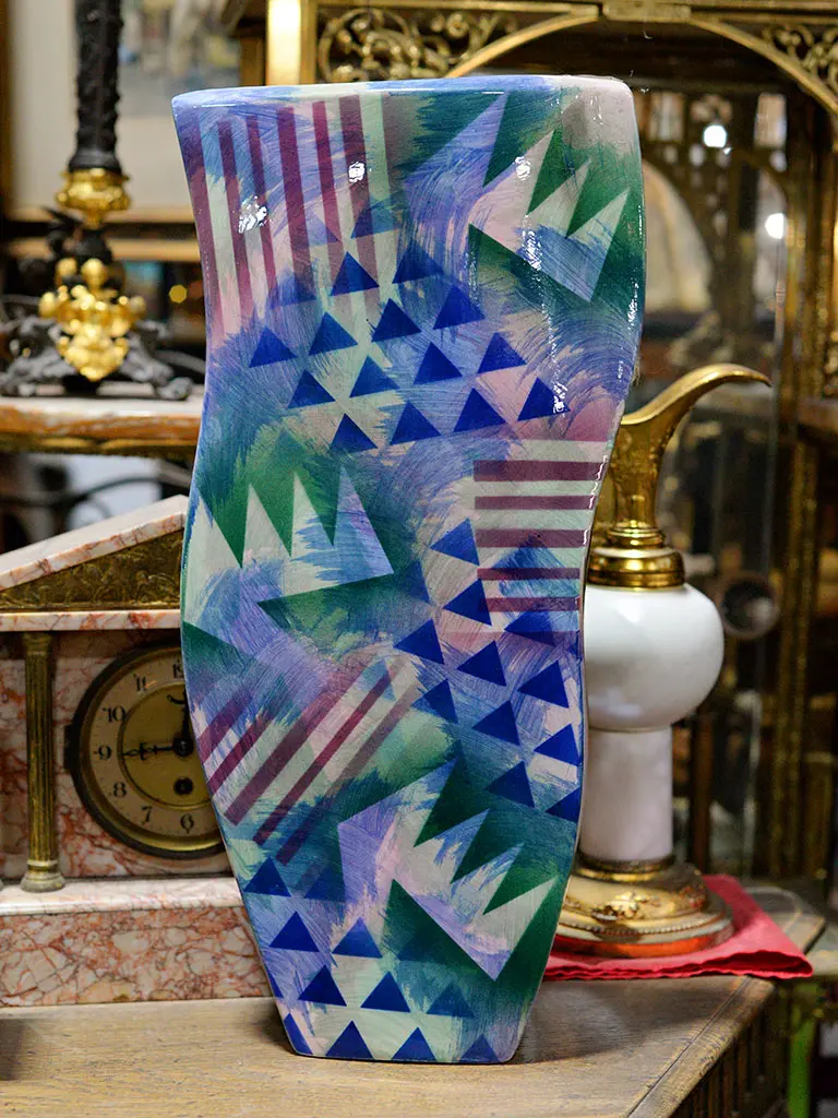 Bold geometric patterned ceramic vase with blue, green and purple triangle motifs displayed at Roadshow Antiques Mall, with an antique marble mantel clock and brass oil lamp visible in the background