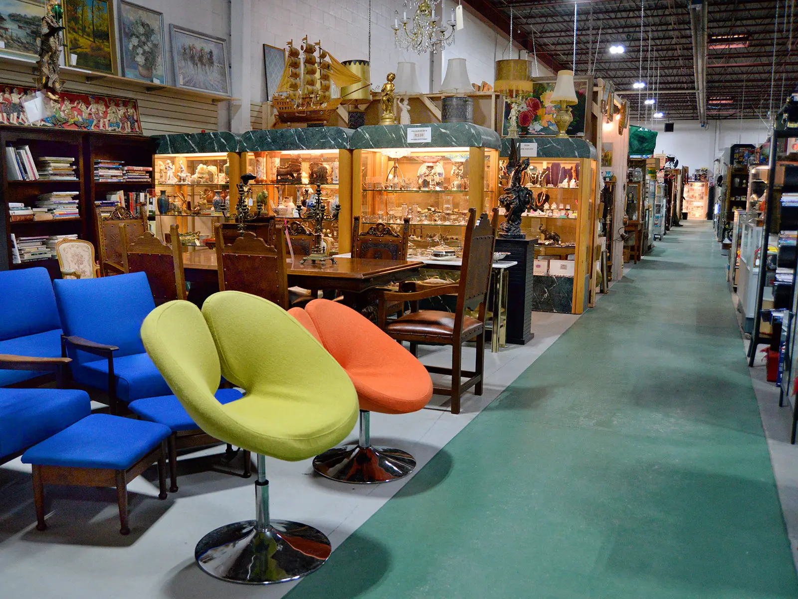Roadshow Antiques Mall aisle showing mid-century modern swivel chairs in lime green and orange, vintage blue armchairs, illuminated display cases filled with collectibles, and rows of vendor booths stretching into the distance