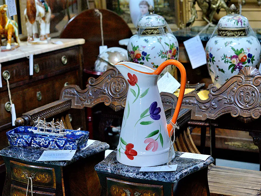 Hand-painted ceramic pitcher with tulip motif displayed on a marble-topped stand at Roadshow Antiques Mall, surrounded by floral ginger jars, ornately carved furniture, and a blue spongeware dish