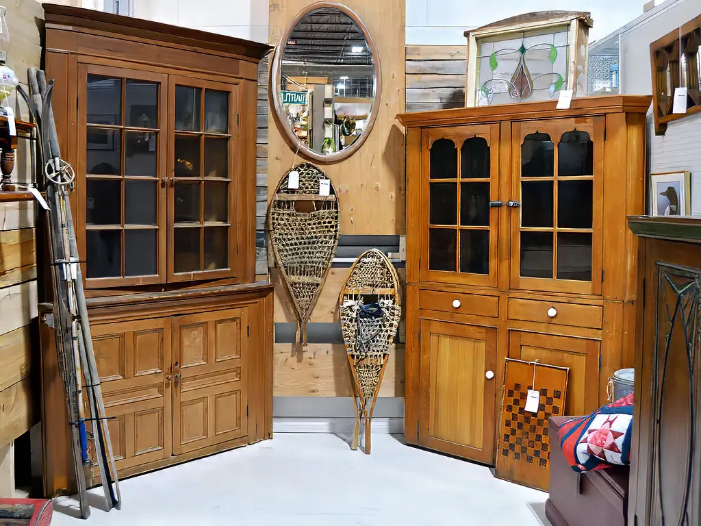 Antique pine corner cabinets and hutches displayed at Roadshow Antiques Mall alongside vintage Canadian snowshoes, an oval mirror, stained glass panel, and a traditional patchwork quilt