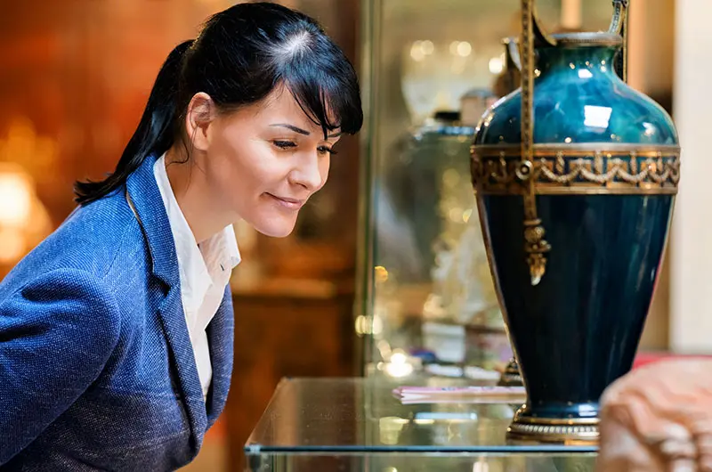 Woman closely examining an ornate teal and gold antique vase in a display case at Roadshow Antiques Mall