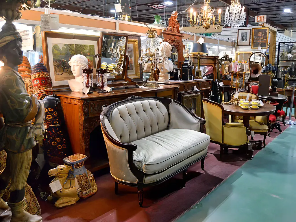 Wide view of Roadshow Antiques Mall showroom featuring a tufted French-style settee, ornate carved desk, marble busts, crystal chandelier, decorative figurines, and an extensive collection of antique furniture and collectibles