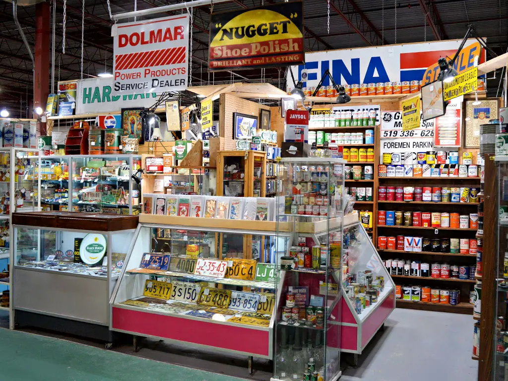 Vintage advertising collectibles booth at Roadshow Antiques Mall featuring antique tin signs, old license plates, retro oil and product tins, and Canadiana memorabilia including Nugget Shoe Polish, Fina, and Dolmar signage