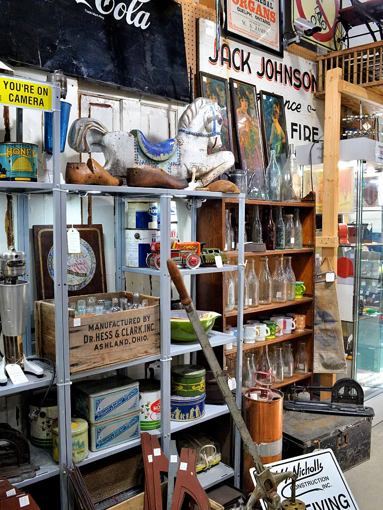 Eclectic Canadiana collectibles booth at Roadshow Antiques Mall featuring a vintage carousel horse, antique wooden crates, old bottles, tin containers, retro signage, and general store memorabilia