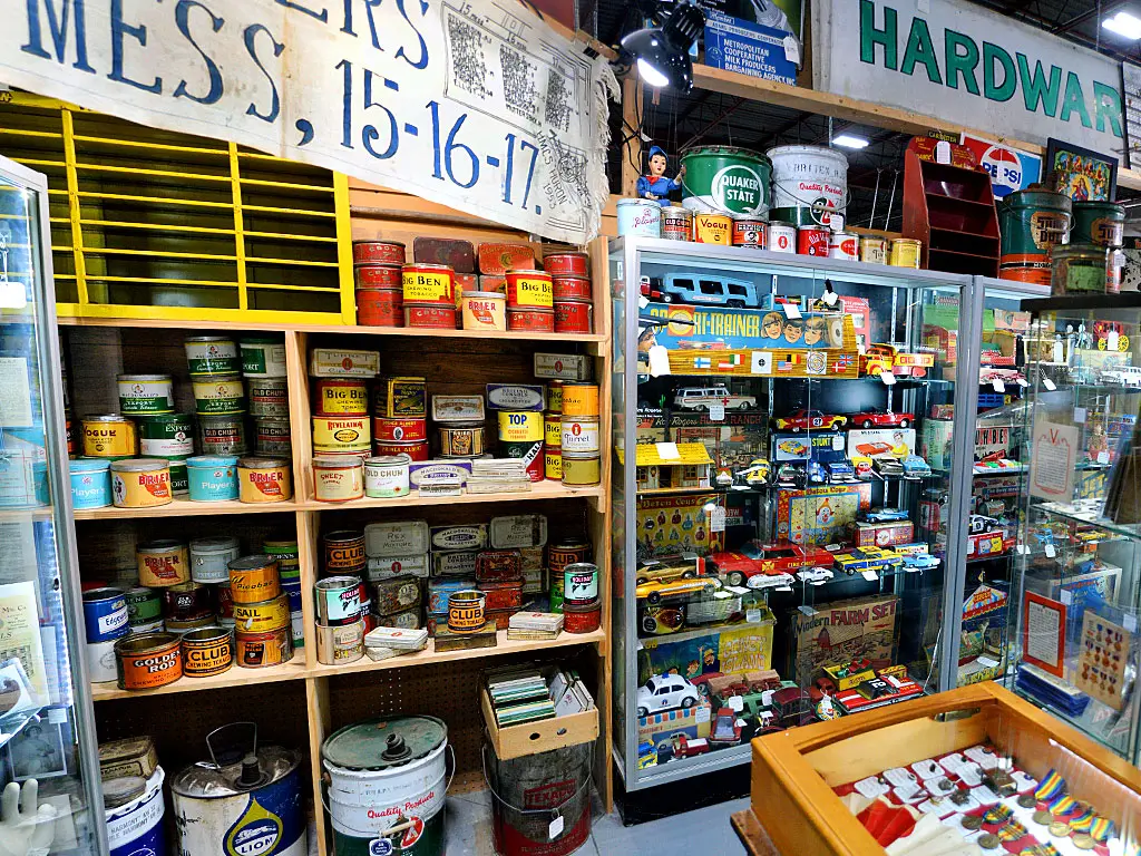 Vintage tobacco tins, advertising cans, and antique toy vehicles displayed in a collector's booth at Roadshow Antiques Mall, with retro signage, die-cast cars, and tin toys filling the shelves